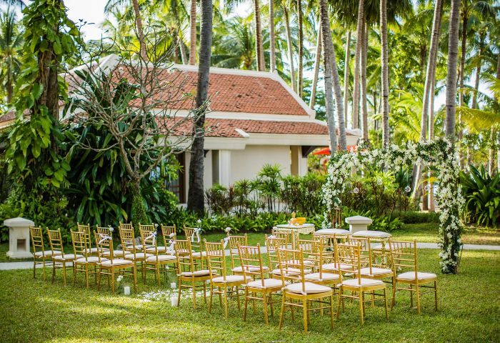 Santiburi Koh Samui wedding seating. Gold chairs face a floral archway on green grass. Palm trees and a building are in the background.