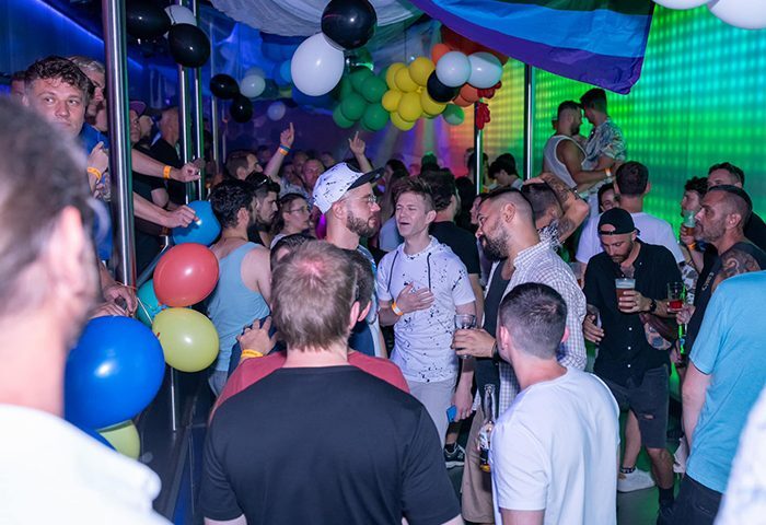 Gay men at Club TerMAX Prague dance under rainbow flag. Balloons, party lights, and drinks create a vibrant atmosphere for gay travel.
