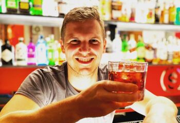The Saints: Gay man at bar holding a cocktail. Travel advice for gay men. The-saints.jpg. Smiling, drinks, nightlife.