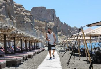 Theros Wave Bar: Man walking on beach at gay-friendly Theros Wave Bar, Santorini. Beach umbrellas and lounge chairs visible. Gay travel.