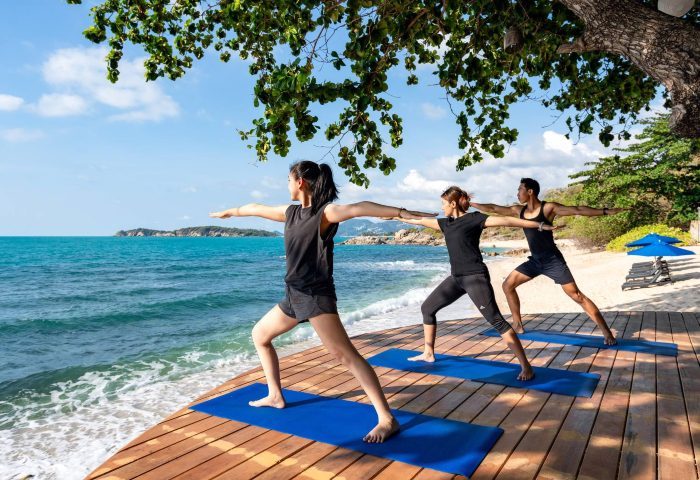 SAii Koh Samui Villas: Yoga session on a wooden deck overlooking the sea. Group of people in warrior pose. Serene tropical beach view.